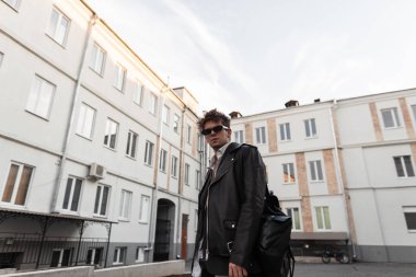 Siyah güneş gözlüklü, modaya uygun siyah ceketli çekici bir genç adam şehirde şık bir sırt çantasıyla poz veriyor. Modern Amerikan hipster adamı. Yeni koleksiyon erkek giyim.