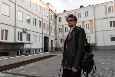 Kentin modern binasının yanında, büyük boy moda siyah deri ceketli, sırt çantalı, modaya uygun genç adam modeli duruyor. Dışarıda güneş gözlüklü yakışıklı hippi..
