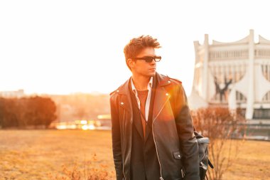 Şık güneş gözlüklü, modaya uygun deri ceketli, antika sırt çantalı modacı genç adam akşamları dışarıda poz veriyor. Çekici hippi adam gün batımında turuncu güneş ışığından hoşlanıyor..
