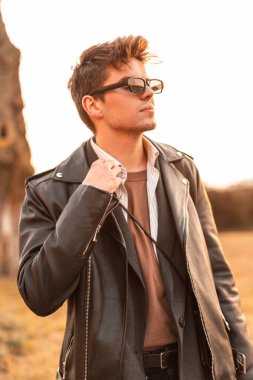 Kaliteli güneş gözlüklü yakışıklı genç hippi siyah ceketli, şık bir saç stili ve doğada parlak turuncu bahar güneşinin tadını çıkarıyor. Çekici adam gün batımında dışarıda dinleniyor..