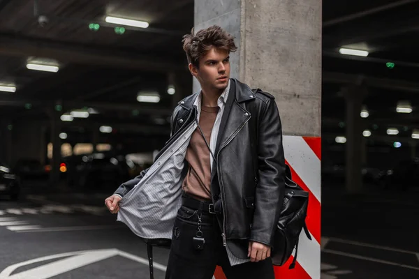 European young man in a fashion black leather jacket in a classic shirt with a vintage backpack with a hairstyle posing near a pillar with a red-white line in a city parking lot. Attractive trendy guy