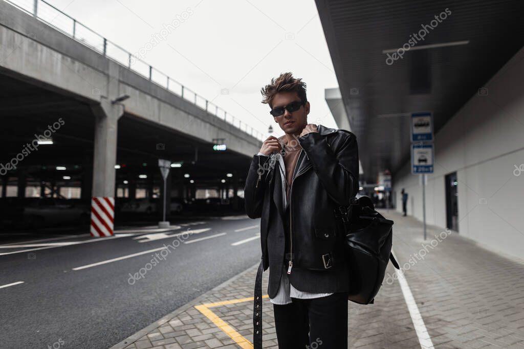 American trendy young man model in a youth oversized fashionable black leather jacket with a stylish hairstyle stands near modern building in the city. Handsome hipster guy in sunglasses in the street