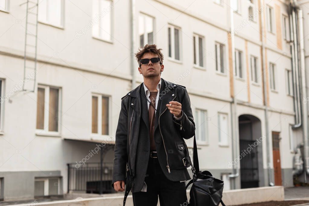 Young man in fashion youth oversized black leather clothes with backpack stand with cigarette near buildings in the city on a spring day. Brutal guy hipster in dark sunglasses enjoy a smoking outdoors