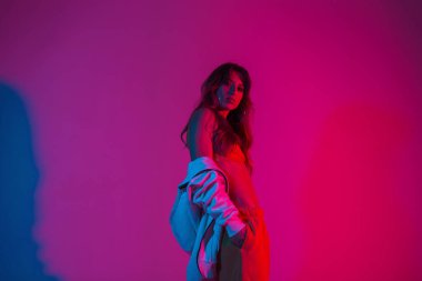 Parlak neon ışıkları olan bir odada kapüşonlu şık giyinmiş şık genç model bir kadın duruyor. Modern moda kız stüdyoda pembe-kırmızı renkli poz veriyor. Moda hanımları. Yaratıcı kavram. 