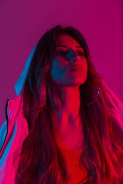 Parlak pembe neon ışığı olan bir odada kapüşonlu uzun saçlı, seksi, modern bir kadın portresi. Şık giyinen güzel bir kız model. Disko tarzında çok renkli giyiniyor..