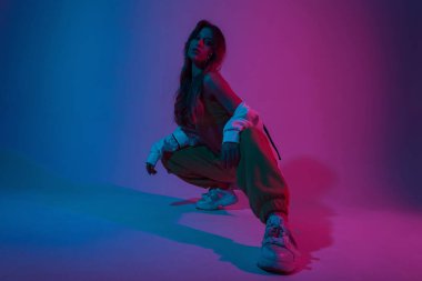 Neon renkli bir odada oturan şık bir spor takımının içinde şık ayakkabılar giyen genç bayan dansçı. Moda kıyafetleri içinde güzel, modern kız model. Pembe, mavi, parlak ışık saçan bir ev..