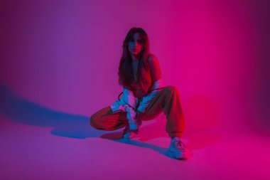 Beyaz spor ayakkabılı modern genç bayan dansçı parlak pembe ışıklı bir odada oturuyor. Stüdyoda renkli ışıkla poz veren güzel bir kız..