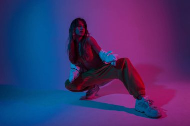 Modern çekici genç bayan dansçı beyaz spor ayakkabılarla şık genç bayan dansçı parlak mavi ışıkla bir odada oturuyor. Stüdyoda neon renkli pozlar veren güzel bir kız model..