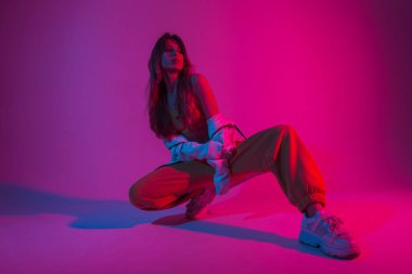 Beyaz spor ayakkabılarıyla şık genç bayan dansçı parlak pembe ışıklarla dolu bir odada oturuyor. Seksi modern kız modeli stüdyoda neon rengi parlak kırmızı pozlar veriyor..