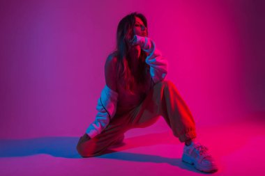 Beyaz spor ayakkabılı, şık, genç ve seksi bir dansçı kız parlak pembe ışıklı bir odada oturuyor. Havalı, güzel bir kız model stüdyoda neon rengi parlak kırmızı renklerle poz veriyor.