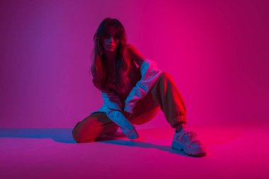 Beyaz spor ayakkabılarıyla şık genç bayan dansçı parlak pembe ışıklarla dolu bir odada oturuyor. Stüdyoda renkli ışıkla poz veren güzel bir kız..