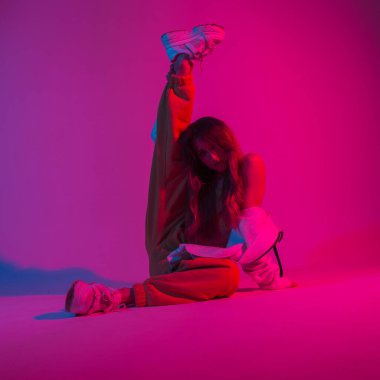 Beyaz spor ayakkabılı seksi genç bayan dansçı parlak pembe ışıklı bir odada oturuyor. Havalı güzel kız modeli stüdyoda neon rengi parlak kırmızı renklerle poz veriyor.