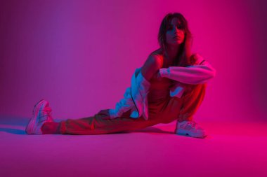 Moda kıyafetleri içinde modern genç bir kadın neon parlak pembe ışıkla kapalı alanda esneme hareketleri yapıyor. Renkli bir stüdyoda poz veren çekici bir kız. Yaratıcı sanat konsepti. Sanat modası.