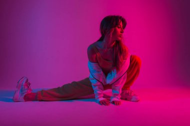 Moda kıyafetleri içinde güzel genç bir kadın neon parlak pembe ışıkla içeride esneme hareketleri yapıyor. Renkli bir stüdyoda poz veren çekici bir kız. Yaratıcı sanat konsepti. Sanat modası.
