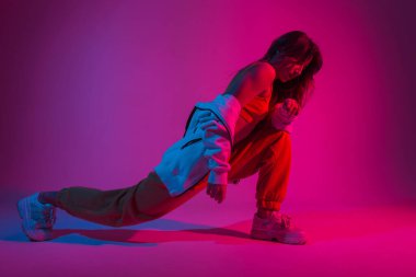 Moda kıyafetleri içinde oldukça modaya uygun genç bir kadın, çok renkli ultraviyole renkli modern bir odada fitness yapıyor. Kız dansçı, stüdyoda pembe ışıkla dans etmeden önce esneme hareketleri yapıyor..