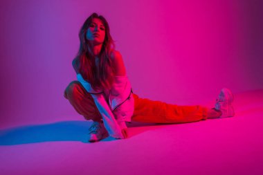 Moda kıyafetleri içinde güzel bir kadın neon parlak kırmızı ışıkla içeride esneme hareketleri yapıyor. Renkli bir stüdyoda poz veren modern bir kız. Yaratıcı sanat konsepti. Sanat modası. Biçim.