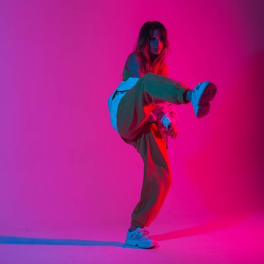 Şık giyinmiş, şık pantolonlu, spor ayakkabılı güzel genç bir dansçı odada tek bacağında parlak pembe morötesi bir renkle poz veriyor. Havalı, şık bir kız, neon renkli ışıkla içeride dans ediyor..