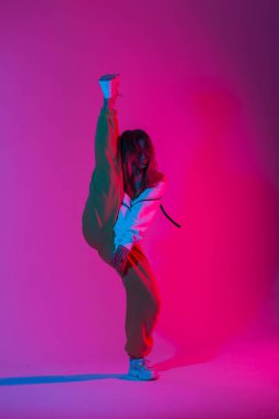 Moda mankeni modern kadın şık kıyafetler içinde şık spor ayakkabıları içinde açık renkli renkte dans ediyor. Parlak neon ışıklı, pembe-mavi bir odada poz veren sportif kız dansçı..