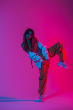 Moda spor ayakkabıları içinde şık genç bayan dansçı stüdyoda parlak pembe bir ışıkla tek ayak üzerinde duruyor. Renkli neon renkli odada dans eden şık bir kız..