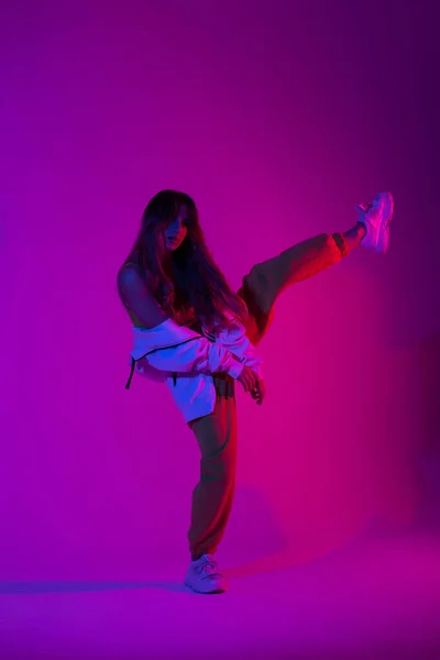 ファッションスニーカーの流行のスポーツの若者の服を着た現代のスタイリッシュな若い女性ダンサーは 明るい青の光でスタジオの1本足に立っています グラマー女の子ダンスでザ部屋でピンクネオン色 — ストック写真