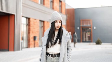 Moda mankeni, şık örgü şapkalı, eski püskü ceketli güzel bir kadın güneşli bir günde modern binanın yanındaki yola bakıyor. Moda kıyafetleri içinde şık bir esmer kız. Sıradan stil.