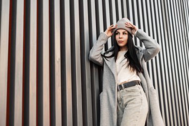 Moda mankeni havalı, esmer, şık bahar kıyafetleri giyen, uzun saçlı, modern metal binanın yanındaki sokakta duran hoş bir kadın. Modern kız şehirde poz veriyor. Kadınlar için moda kıyafetler.