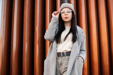 Şehirde metal duvarın yanında poz veren şık montlu, eski moda gözlüklü, gri şapkalı şık, genç hippi bir kadın. Sokaktaki Avrupalı modaya uygun kız modeli. Bahar gündelik tarzı