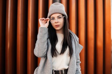 Moda kıyafetleri içinde genç ve güzel bir hippi kadın bardakları düzeltiyor. Moda portresi bej kazaklı güzel kız modeli örülmüş moda şapkalı metal binanın yanındaki gri moda ceketli..
