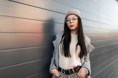 Moda gözlüklü, modaya uygun örülmüş şapkalı, modaya uygun genç ve çekici bir kadın şehrin dışında gri metal duvarın yanında duruyor. Güzel hippi kız sokakta poz verir..