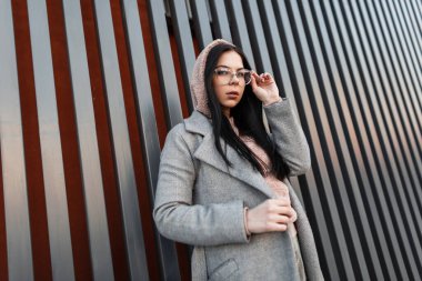 Şık, şık, uzun gri montlu hippi genç bayan şehirdeki klasik metal duvarın yanında moda gözlüklerini düzeltiyor. Modern moda hippi kız dışarıda poz veriyor. Sokak havası..