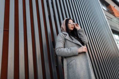 Moda mankeni, modaya uygun, uzun gri montlu hippi bir kadın şehirde klasik metal duvarın yanında gözlük takıyor. Modayı takip eden hippi kız dışarıda poz veriyor. Modern bahar gündelik görünümü.