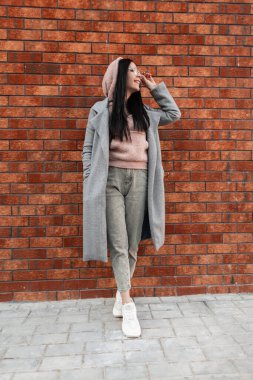 Moda tutkunu genç bayan hippi, şık gençlik gündelik kıyafetleriyle mutlu bir şekilde gülümsüyor, şık spor ayakkabılar giyiyor, klasik tuğla duvarın yanında gözlüklü, açık havada. Tatlı pozitif kız poz veriyor ve sokakta gülümsüyor..