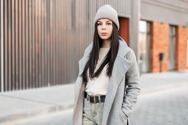 灰色のコートでベージュのセーターのニット帽のかなりスタイリッシュな若いブルネットの女性は 明るい晴れた日にヴィンテージの建物の近くの通りを歩く カジュアルウェアで美しい女の子のファッションモデル ストリートスタイル — ストック写真