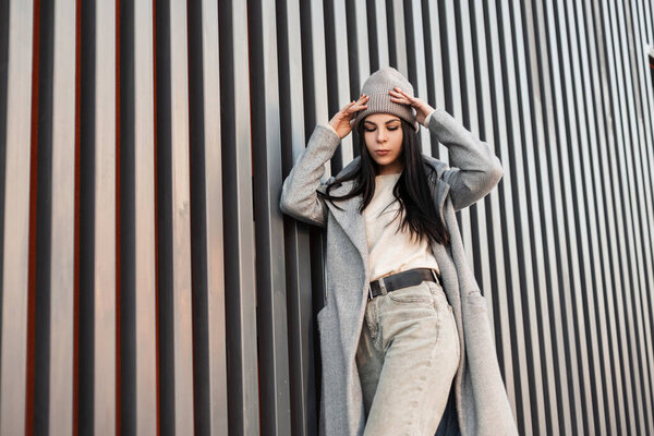 European young brunette woman with long hair in stylish gray coat in jeans in knitted trendy hat posing near vintage metal wall. Pretty attractive girl fashion model in spring clothes. Casual look.