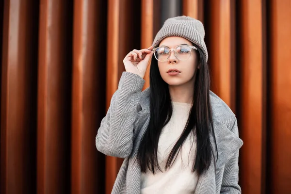 Şık, kentli, güzel, esmer, modaya uygun örülmüş gri şapkalı, sokakta klasik ceketli bir kadın portresi. Avrupalı modacı hippi kız moda gözlüklerini düzeltiyor. Sıradan bahar modası.