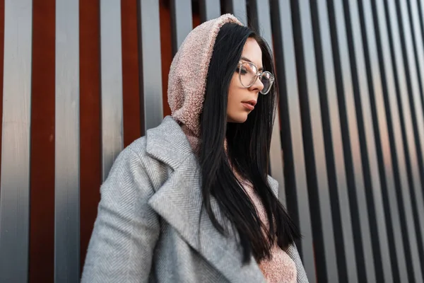 Urban cute young hipster woman with black long hair in trendy pink hoodie in stylish gray coat in fashionable glasses stands and looking down near building in the city. Cool girl model. Youth style.