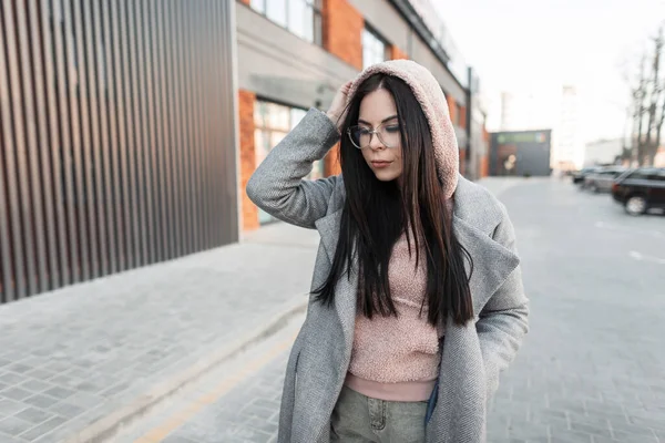 Attractive cute young hipster woman with black long hair in trendy pink hoodie in stylish gray coat in fashionable glasses poses and looking down near building in the city. Urban girl on the street.