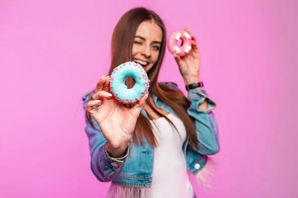 Beautiful funny young woman model with cute smile in stylish jeans clothes holds donuts in sweet glaze in hands on a pink background indoors. Cheerful girl poses with tasty desserts. Lady with sweets.