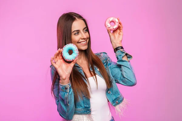 Trendy young funny woman model in stylish white t-shirt with fashionable blue denim jacket posing with donuts in sweet glaze near the pink wall in the room. Positive cheerful girl in the studio.