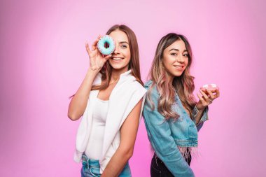 Şık kot elbiseli komik kız arkadaş kadınlar odadaki klasik duvarda pembe yüzün yanında tatlı renkli çörekler tutuyorlar. Tatlılarla poz veren ve gülümseyen güzel kızlar..