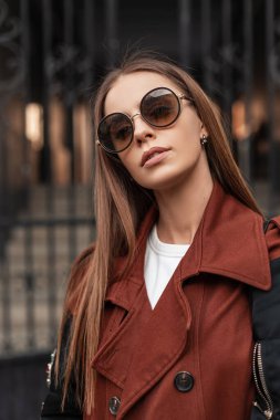 Moda portresi, modaya uygun, siyah gözlüklü, demir kapının yanında uzun saçlı, trençkotlu güzel hippi kadın. Avrupalı modern kız model açık havada. Sıradan stil