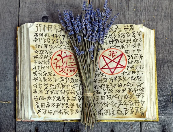 Old book with black magic symbols and lavender bunch on witch table. Occult, esoteric, divination and wicca concept. Halloween vintage background. No foreign text, all symbols on pages are fantasy, imaginary ones.