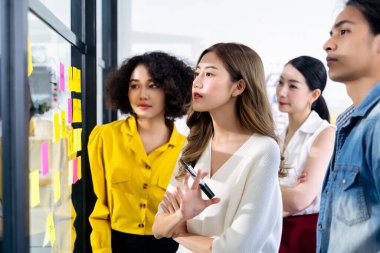 Bir grup Asyalı iş adamı modern ofiste buluşuyor. Genç yaratıcı ekip cam duvardaki yapışkan notu paylaşmak için post it notlarını kullanıyor. ve tasarım planlaması ve beyin fırtınası odaklama konsepti. 