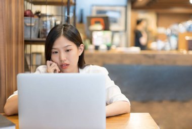 Mutlu genç Asyalı kadın telefon kullanıyor ve kafeterya ofisinde dizüstü bilgisayarla çalışıyor..