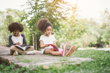 Küçük Afro kız yeşil çiviler arasında kitap okuyor arkadaşlarıyla çayır bahçesinde eğitim kavramını okuyor.
