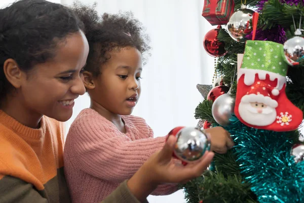 Anne ve kızı Afrikalı Amerikalı kız Noel ağacını süslüyor, Noel ışıklarını ayarlıyor ve eğleniyorlar.