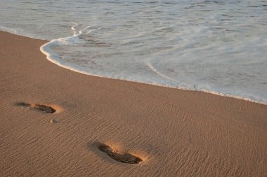 Gün batımında dalganın köpüğünün yanında kumda bir adamın ayak izleri.