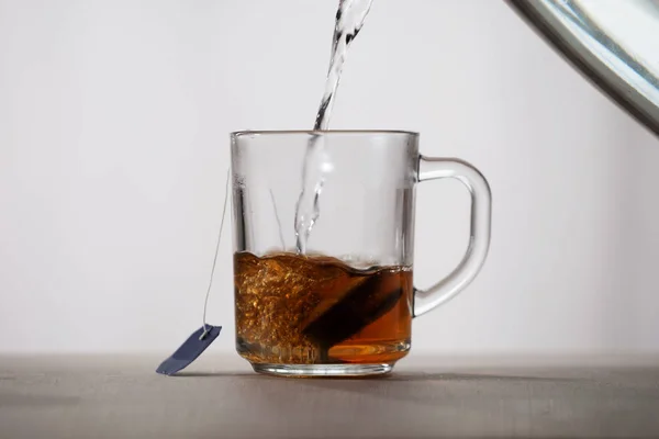 Boiling water is poured into a glass mug with a tea bag. Water turns into tea, it is painted in amber color. Front view