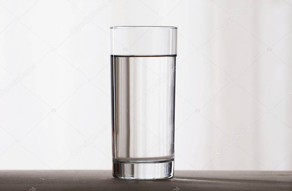 A simple tall glass filled with clear water stands on a wooden surface on a light background. Front view