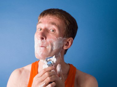 Yakın plan. Adam mavi bir arka planda çenesini heyecanla tıraş ediyor. Mavi arkaplanda stüdyo fotoğrafçılığı